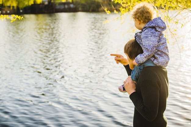 Photo gratuite père qui pointe le lac avec son fils sur ses épaules