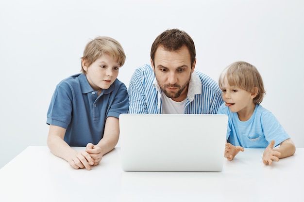 Père regardant des dessins animés avec des fils. Portrait de père attrayant enthousiaste avec de jeunes garçons assis à table et regardant l'écran du portable avec intérêt et curiosité