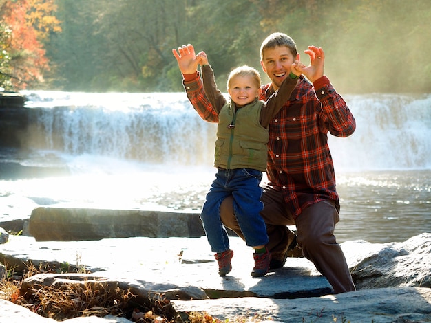 Père souriant avec son fils dans un parc entouré de verdure et une cascade sous la lumière du soleil