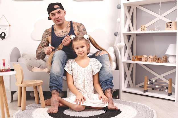 Père tatoué de temps drôle dans une casquette et son enfant jouent à la maison papa fait les cheveux de ses filles...