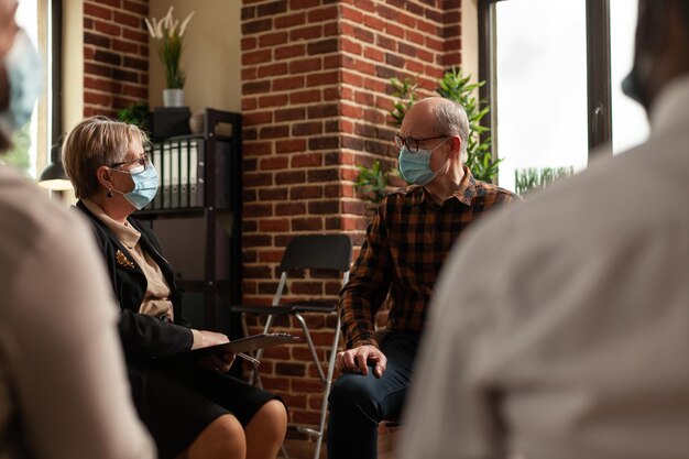 Personne âgée avec masque facial parlant aux patients en cercle lors d'une réunion, pour guérir la dépendance à l'alcool lors d'une thérapie de groupe de soutien. Homme discutant avec un psychologue et des gens de problèmes.