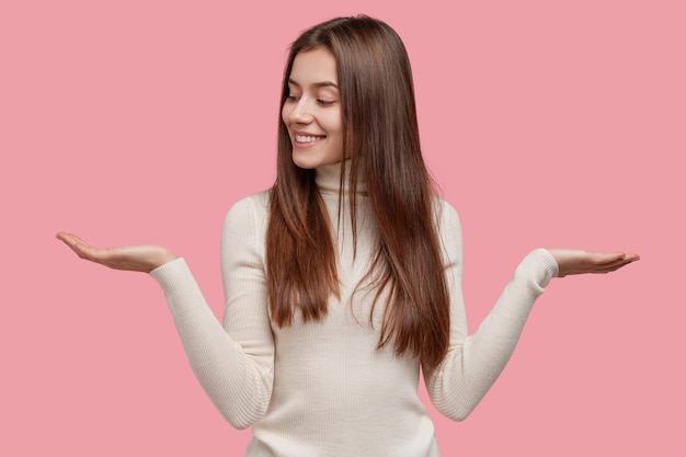 Une personne heureuse lève deux paumes, sent l'hésitation entre deux articles, a une expression faciale ravie, vêtue d'un pull blanc chaud