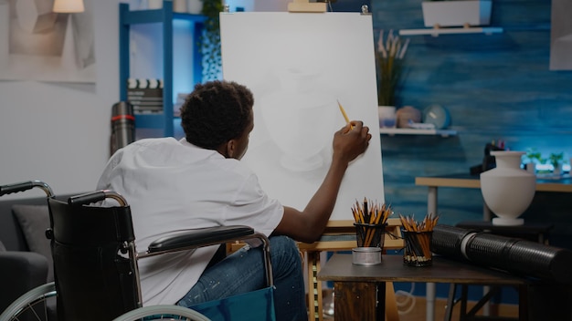 Personne noire assise dans un vase de dessin d'espace d'œuvres d'art à l'aide d'une toile blanche et d'un crayon de table. Artiste afro-américain handicapé en fauteuil roulant créant un design comme passe-temps artistique