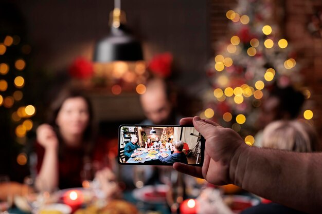 La personne prend des photos de personnes à table en train de profiter d'un dîner de fête avec des verres d'alcool entourés d'un arbre de Noël et d'ornements. Divers amis et famille posant pour des photos la veille de Noël.