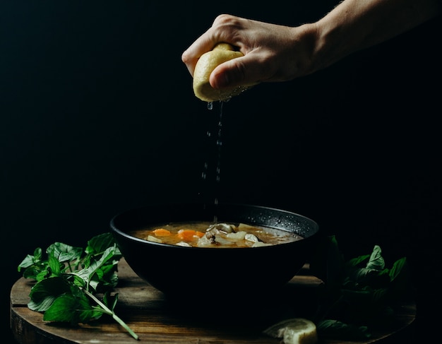 Personne pressant le citron sur la soupe dans un bol noir avec un mur sombre