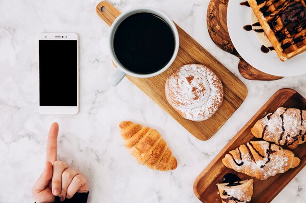 Une personne qui pointe du doigt un téléphone portable et des petits pains cuits au four; croissant; gaufres et tasse à café sur le bureau