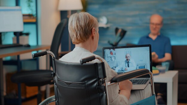 Personne retraitée en fauteuil roulant utilisant un appel vidéoconférence pour rencontrer un médecin pour une consultation en ligne sur ordinateur portable. Vieille femme handicapée physique parlant à un médecin lors d'une conférence de télésanté.