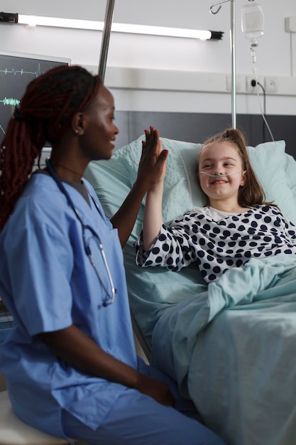 Le personnel de l'établissement de soins de santé à l'enfance élève un enfant malade sous traitement médical. Infirmière faisant cinq gestes avec une fille malade se reposant dans le lit du patient du service pédiatrique de l'hôpital.
