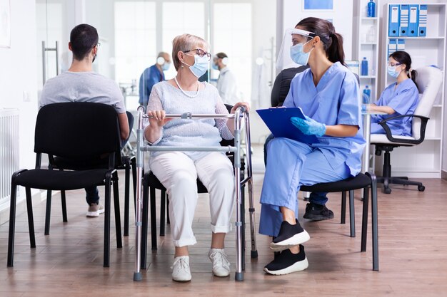 Personnel médical discutant du traitement avec une femme âgée handicapée portant un déambulateur dans la salle d'attente de l'hôpital portant un masque facial contre le coronavirus