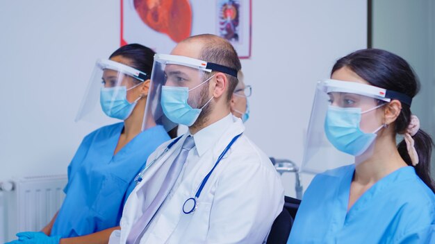 Personnel médical avec masque facial et visière contre le coronavirus assis sur des chaises dans la salle d'attente de l'hôpital. Medic portant un stéthoscope.