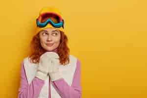 Photo gratuite personnes, loisirs, pensées, concept d'activités de loisirs. adorable femme au gingembre garde les mains jointes sur la poitrine, porte une tenue chaude, un masque de snowboard, pense à une nouvelle aventure en hiver