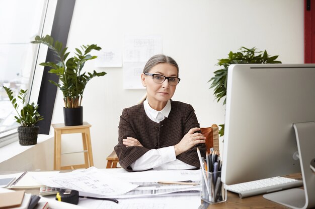Personnes, profession, emploi et âge. Architecte de femme senior professionnelle confiante dans des vêtements formels et des lunettes assis sur son lieu de travail, les bras croisés, faire des dessins et utiliser le programme de CAO de pc