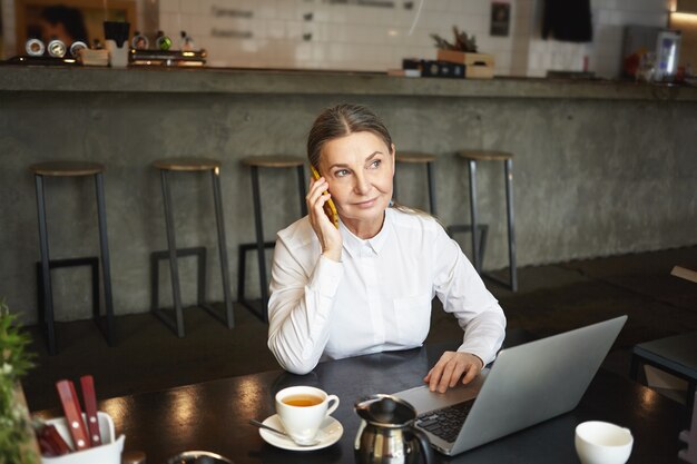 Personnes, travail, style de vie moderne, technologie et concept de communication. Photo d'une femme d'affaires mature confiante attrayante en tenue de soirée ayant un appel téléphonique pendant une pause-café au café et à l'aide d'un ordinateur portable