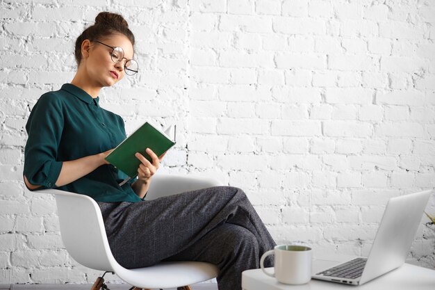 Personnes, travail, technologie et concept de mode de vie moderne. Portrait de femme d'affaires sérieuse concentrée dans des lunettes élégantes travaillant à distance au café, écrit dans le journal, assis avec un ordinateur portable et une tasse