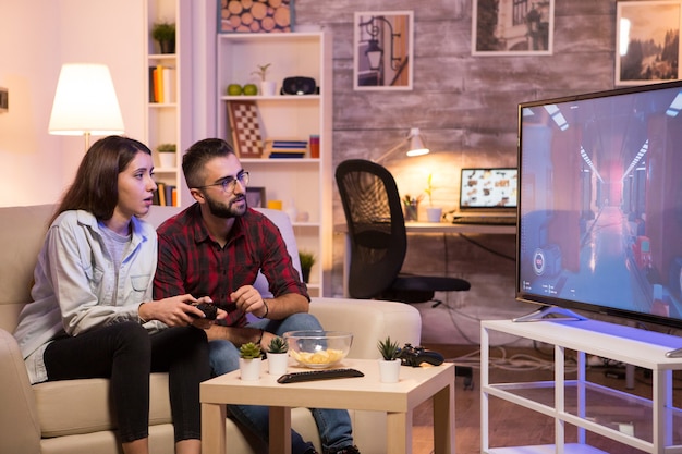 Petit ami apprenant à sa petite amie à jouer à des jeux vidéo à la télévision à l'aide de contrôleurs. Couple assis sur le canapé.