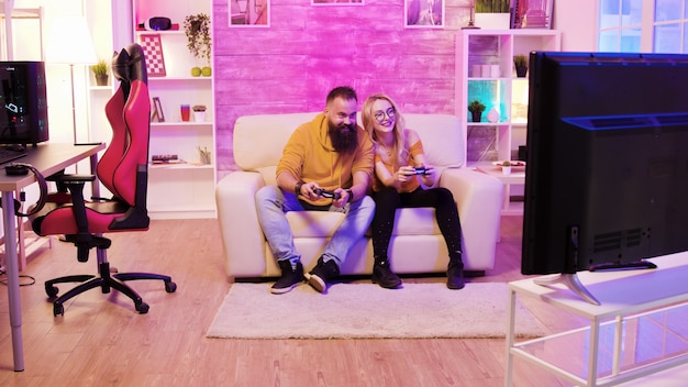 Petit ami barbu jouant à des jeux en ligne avec sa belle petite amie blonde assise sur leur canapé à l'aide de contrôleurs sans fil.