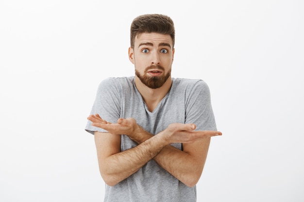 Petit ami confus ne peut pas comprendre ce qui a mal fait. Désemparé et interrogé charmant brunet avec barbe et yeux bleus haussant les épaules avec les bras croisés ouvrant la bouche debout troublé sur le mur gris