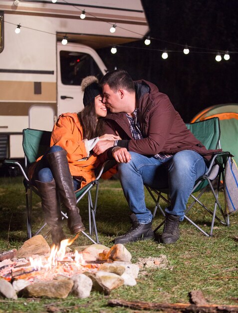 Petit ami embrassant sa petite amie près d'un feu de camp chaud dans une froide nuit d'automne dans les montagnes avec un camping-car rétro en arrière-plan.