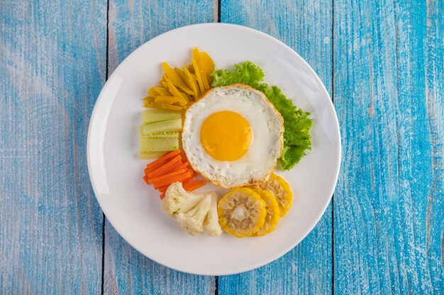 Petit déjeuner américain sur une table bleue avec œuf au plat, salade, citrouille, concombre, carotte, maïs, chou-fleur et tomate.