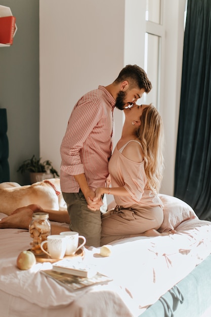 Petit déjeuner au lit. Couple tient la main et embrasse, se regardant dans les yeux dans leur confortable chambre lumineuse.