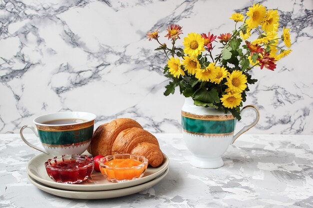 Petit déjeuner français avec croissants, confiture d'abricot, confiture de cerise et une tasse de thé, fleurs rouges et jaunes