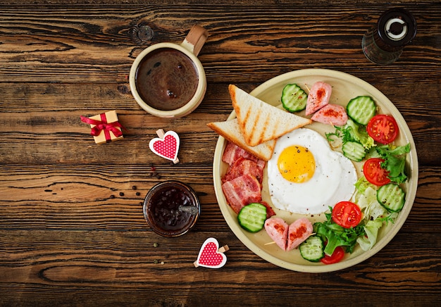 Petit déjeuner le jour de la Saint-Valentin - œuf au plat en forme de coeur, toasts, saucisse, bacond de légumes frais. Petit déjeuner anglais. Tasse de café. Vue de dessus