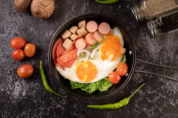 Petit-déjeuner avec des œufs au plat, des saucisses et du jambon dans une poêle avec des tomates. Chili et basilic