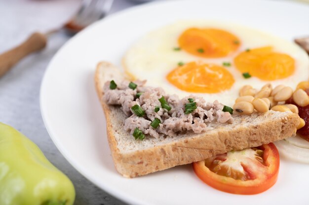 Le petit-déjeuner se compose d'œufs au plat, de saucisses, de porc haché, de pain, de haricots rouges et de soja sur une assiette blanche.