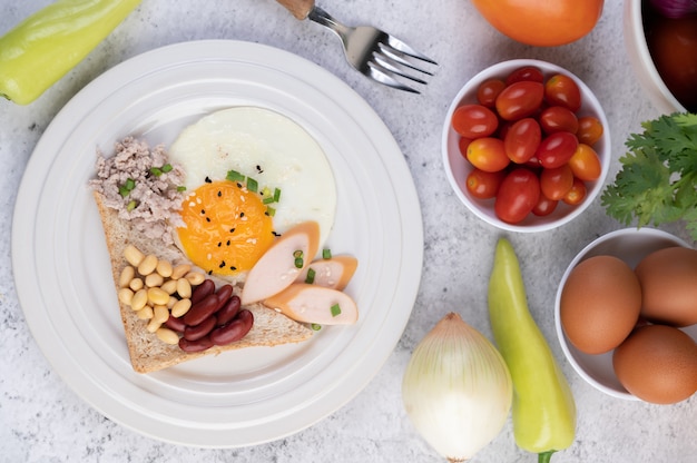 Le petit-déjeuner se compose d'œufs au plat, de saucisses, de porc haché, de pain, de haricots rouges et de soja sur une assiette blanche.