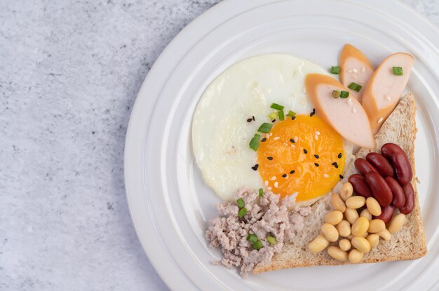 Le petit-déjeuner se compose d'œufs au plat, de saucisses, de porc haché, de pain, de haricots rouges et de soja sur une assiette blanche.