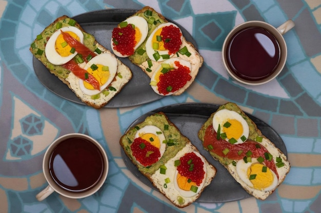 Petit-déjeuner avec thé et sandwichs à l'avocat, fromage à la crème, œufs en tranches, caviar rouge et saumon salé décoré d'oignon vert