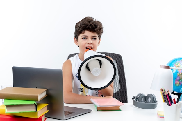 petit écolier assis au bureau avec des outils scolaires parle sur haut-parleur