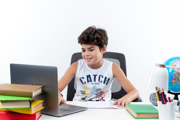 Petit écolier assis au bureau avec des outils scolaires utilisé ordinateur portable isolé sur blanc