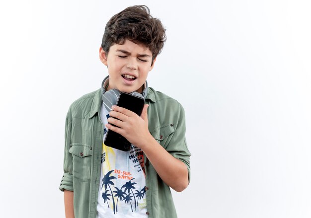 Petit écolier portant des écouteurs tenant le téléphone et chante isolé sur blanc