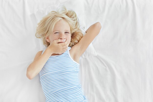 Petit enfant aux cheveux blonds, aux yeux bleus et à la peau tachetée de rousseur, couché dans son lit, couvrant la bouche avec la main et bâillant. Adorable petite fille se réveillant le matin, ayant une expression endormie après le sommeil