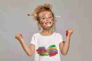 Photo gratuite petit enfant ayant un pinceau dans sa jolie coupe de cheveux, vêtu d'un t-shirt blanc enduit. elle pose avec des mains et des joues peintes, fait des grimaces et regarde la caméra, sur un fond gris.