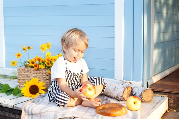 Petit garçon blond assis sur un porche en bois à la maison et mange une pomme enfant heureux et fruits