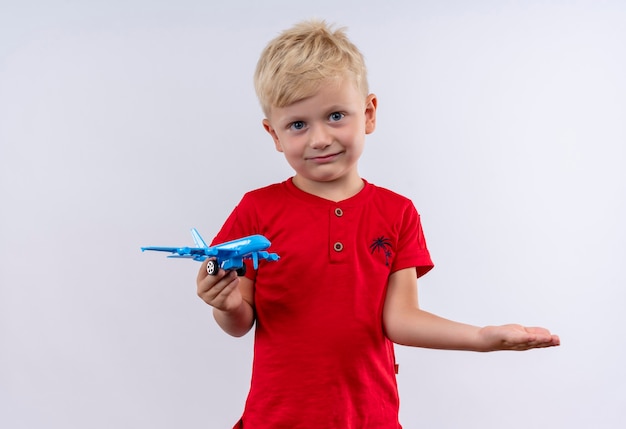 Un petit garçon blond mignon en t-shirt rouge tenant un avion jouet bleu tout en regardant sur un mur blanc