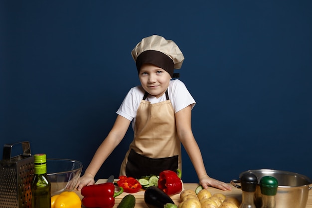 Petit garçon chef en couvre-chef et tablier préparant des aliments sains, regardant et souriant à la caméra tout en se tenant à la table de la cuisine, coupant des légumes pour le dîner. Enfance, cuisine et végétarisme