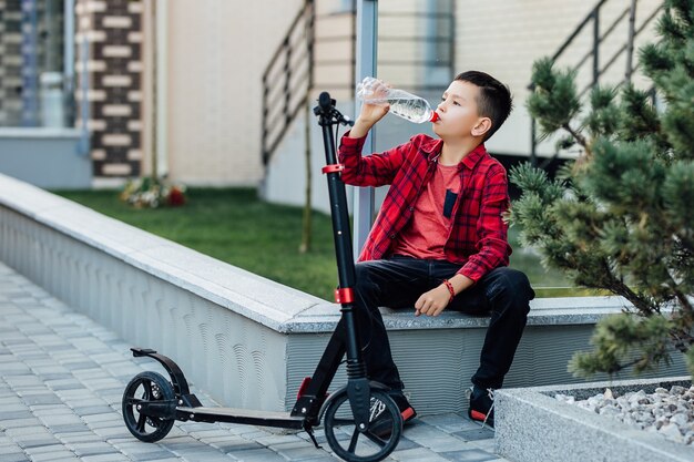 Petit garçon en chemise rouge décontractée assis près de son scooter et de l'eau potable.
