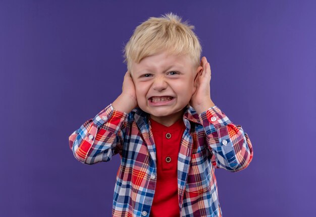 Un petit garçon en colère aux cheveux blonds portant une chemise à carreaux en gardant les mains sur les oreilles