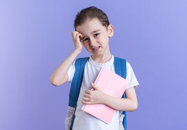 Petit garçon confus portant un sac à dos tenant un bloc-notes regardant la caméra touchant la tête isolée sur un mur violet