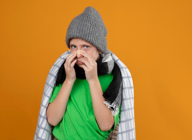 Petit garçon malade portant un chapeau chaud et une écharpe enveloppée dans une couverture mettant un patch sur son nez malheureux et malade debout sur un mur orange