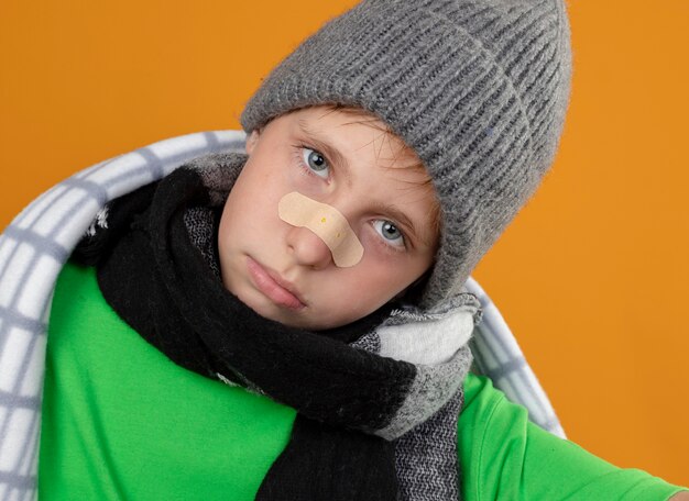 Petit garçon malade portant un chapeau chaud et une écharpe enveloppée dans une couverture avec patch sur son nez malheureux et malade regardant la caméra debout sur fond orange