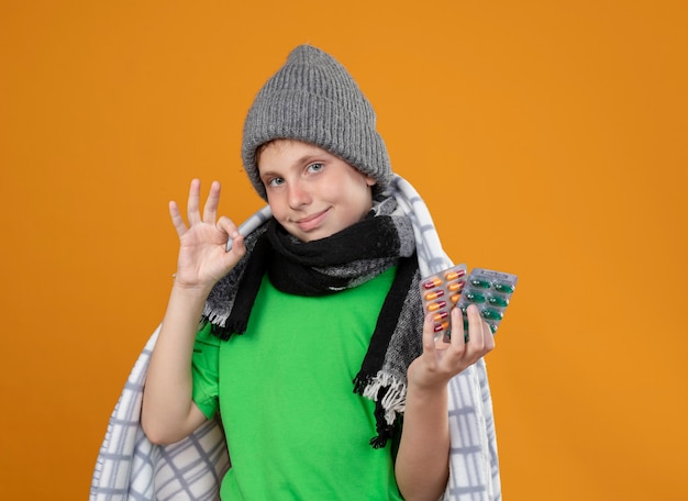 Petit garçon malade portant un chapeau chaud et une écharpe enveloppée dans des couvertures montrant des pilules se sentir mieux à sourire montrant ok chanter debout sur un mur orange