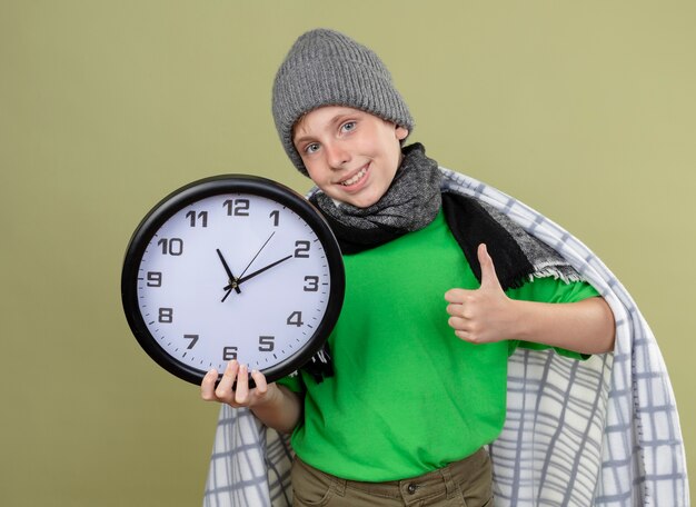 Petit garçon malade portant un t-shirt vert dans une écharpe chaude et un chapeau enveloppé dans une couverture tenant une horloge murale souriant montrant les pouces vers le haut se sentant mieux debout sur un mur léger