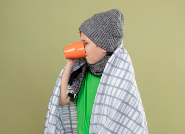 Petit garçon malade portant un t-shirt vert en écharpe chaude et un chapeau enveloppé dans une couverture de boire du thé chaud souffrant de froid debout sur un mur léger