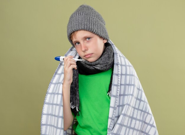 Petit garçon malade portant un t-shirt vert en écharpe chaude et un chapeau enveloppé dans une couverture montrant thermomètre malheureux et malade debout sur un mur léger