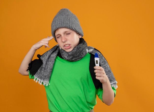 Photo gratuite petit garçon malade portant un t-shirt vert en écharpe chaude et un chapeau montrant un thermomètre se sentant malade et malheureux faisant un geste de pistolet près de temple debout sur un mur orange