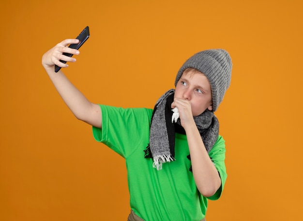 Petit garçon malade portant un t-shirt vert en écharpe chaude et un chapeau se sentant mal tenant un smartphone faisant selfie tout en toussant debout sur un mur orange